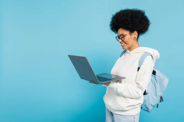 Cheerful african american student with backpack looking at laptop isolated on blue - foto de stock