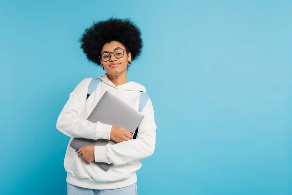 Усміхнений афроамериканський студент у білому светрі та окулярах тримає ноутбук, дивлячись ізольовано на синьому — стокове фото