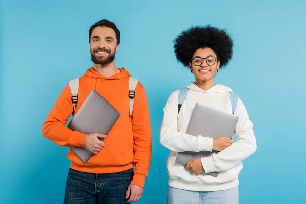 Cheerful interracial couple of students holding laptops and looking at camera isolated on blue - foto de stock