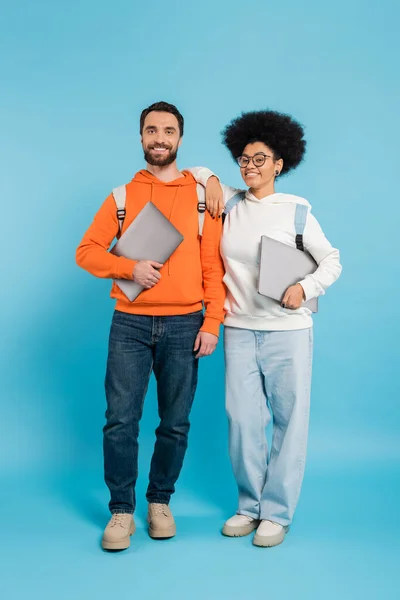 Full length of happy and stylish multiethnic students with laptops smiling at camera on blue - foto de stock