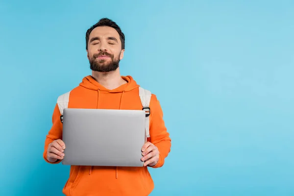 Positive student in orange hoodie holding laptop while standing with closed eyes isolated on blue — Stock Photo