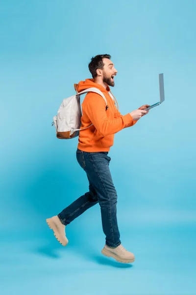 Side view of excited student with backpack and laptop levitating on blue background — Stockfoto