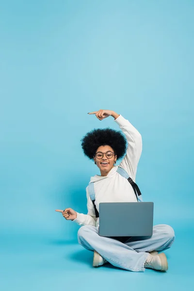 Full length of smiling african american student sitting with laptop and pointing with fingers on blue background - foto de stock