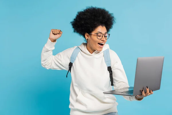 Excited african american student in eyeglasses looking at laptop and showing win gesture isolated on blue — Stock Photo