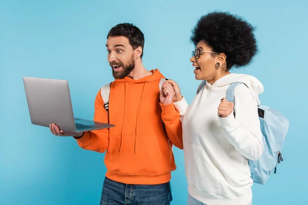 Excited interracial students in hoodies looking at laptop isolated on blue — Stockfoto