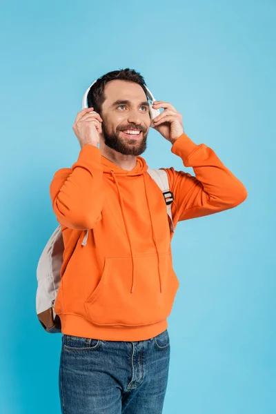 Happy bearded student in orange hoodie adjusting wireless headphones isolated on blue — Stock Photo