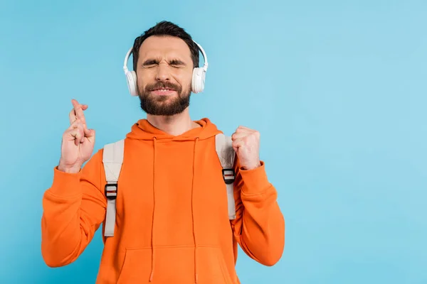 Positive bearded student in wireless headphones crossing fingers while standing with closed eyes isolated on blue — Stock Photo