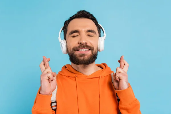 Positive student in wireless headphones holding crossed fingers while standing with closed eyes isolated on blue — Fotografia de Stock