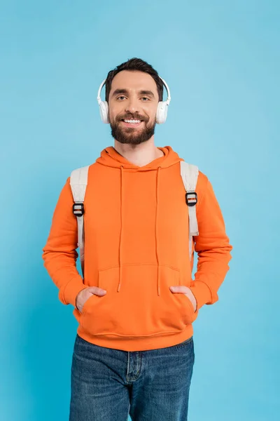 Pleased student in wireless headphones and orange hoodie posing with hands in pockets isolated on blue — Stock Photo