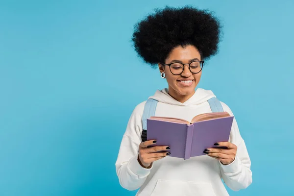 Хвилястий і стильний африканський студент в окулярах посміхається, читаючи книжку ізольовано на синьому — стокове фото