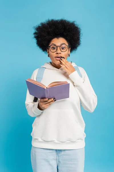 Surprised african american student in eyeglasses standing with book and holding hand near mouth isolated on blue — Photo de stock