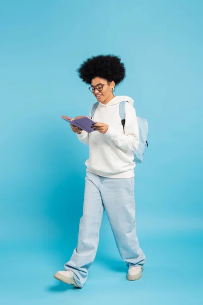 Full length of smiling african american student in eyeglasses and jeans reading book while walking on blue - foto de stock