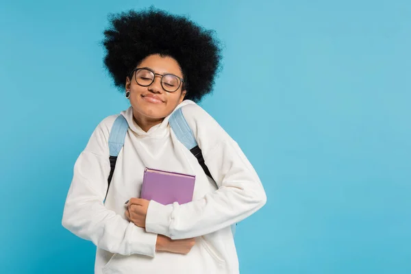 Задоволений афроамериканською студенткою в окулярах, яка тримає книжку, посміхаючись із заплющеними очима, ізольованими на синьому — стокове фото