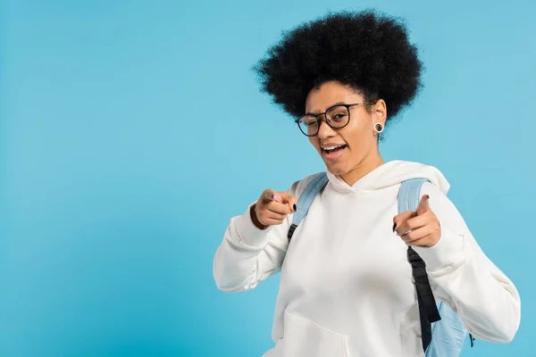 Positive african american student in eyeglasses and white hoodie pointing with fingers and looking at camera isolated on blue — Stock Photo