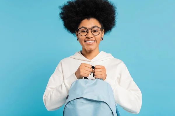 Happy african american student in white hoodie and eyeglasses holding backpack isolated on blue — Stockfoto