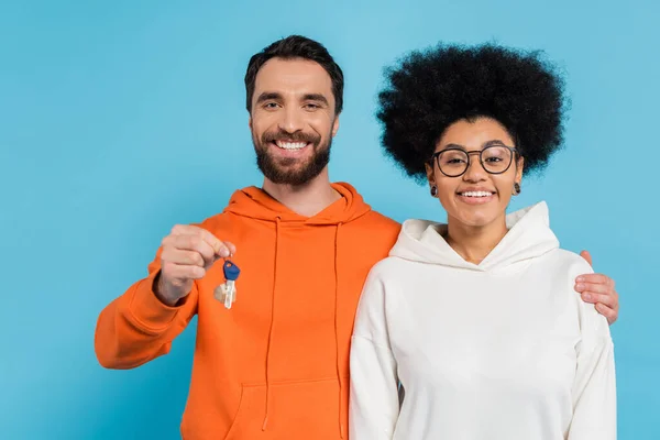Joyful bearded man hugging african american girlfriend and showing key isolated on blue — Stock Photo