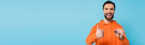 Excited brunette man in orange hoodie showing like gesture while holding key isolated on blue, banner — Photo de stock