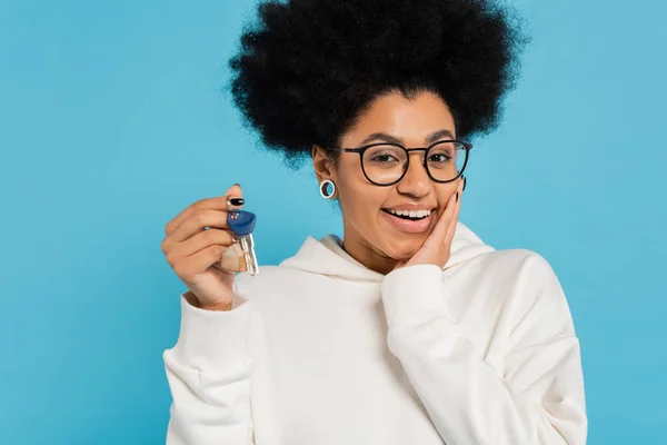 Happy african american woman in stylish eyeglasses and white hoodie touching face while holding key isolated on blue — Stock Photo