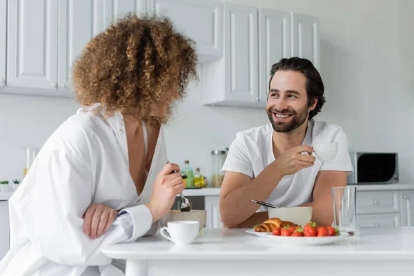 Cheerful bearded man smiling while looking at curly girlfriend during breakfast — Fotografia de Stock