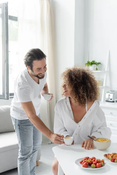 Happy man giving cup of coffee to young curly girlfriend during breakfast in kitchen — Foto stock