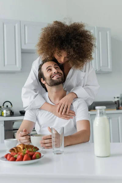 Curly young woman hugging cheerful boyfriend during breakfast - foto de stock