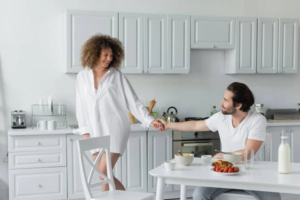 Bearded man holding hands with cheerful woman during breakfast in kitchen — Photo de stock