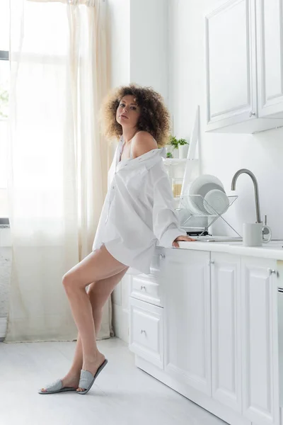 Sexy junge Frau im weißen Hemd steht neben der Arbeitsplatte in der Küche und blickt in die Kamera — Stockfoto