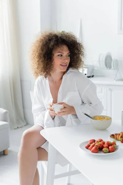 Curly young woman in white shirt holding cup of coffee near tasty breakfast on table — Photo de stock