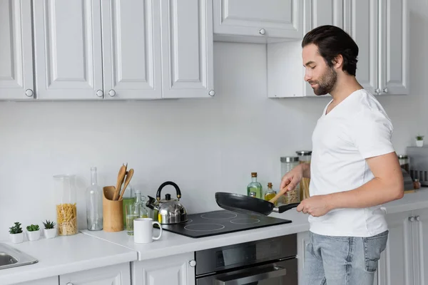 Bearded man in jeans and white t-shirt making breakfast in kitchen - foto de stock