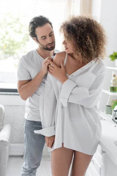 Bearded man undressing curly and sensual woman in white shirt in kitchen — Photo de stock