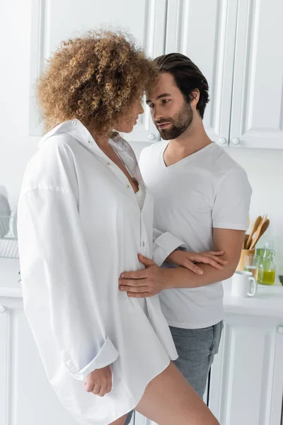 Jeune homme en jeans étreignant femme sensuelle en chemise blanche dans la cuisine — Photo de stock