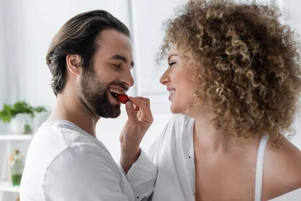 Curly woman feeding cheerful boyfriend with fresh strawberry — Stock Photo