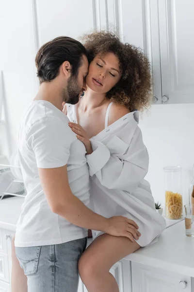 Bearded man in jeans seducing curly young woman with closed eyes sitting on kitchen worktop - foto de stock