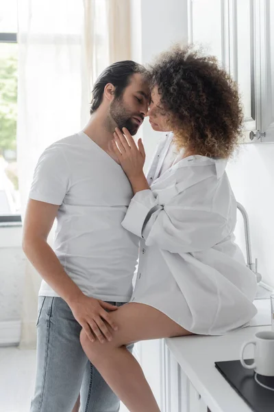 Bearded man in jeans seducing curly young woman in shirt sitting on kitchen worktop — Stockfoto