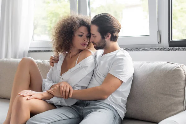 Passionate man sitting on couch and hugging sensual woman in white shirt — Photo de stock