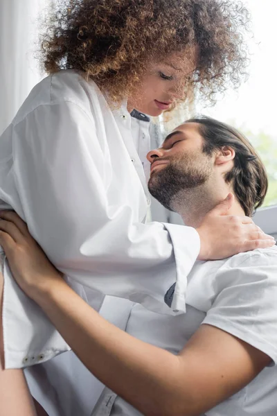 Curly young woman in white shirt hugging bearded man with closed eyes - foto de stock