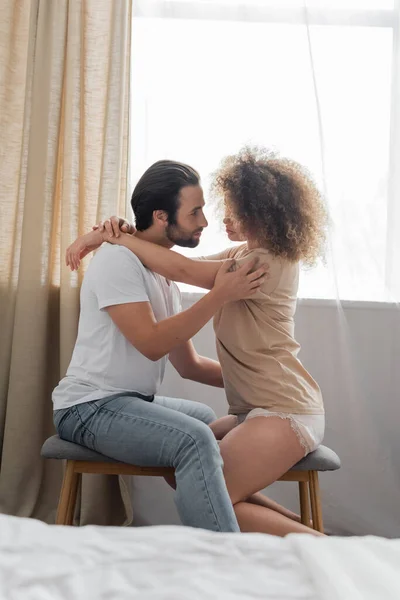 Side view of bearded man sitting on bed bench and hugging sexy girlfriend in bedroom - foto de stock