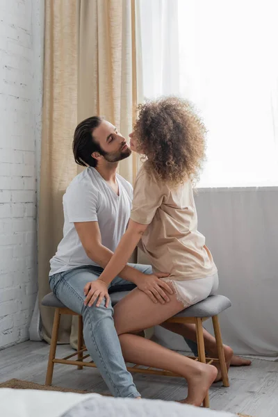 Bearded man sitting on bed bench while hugging seductive girlfriend in bedroom — Stock Photo