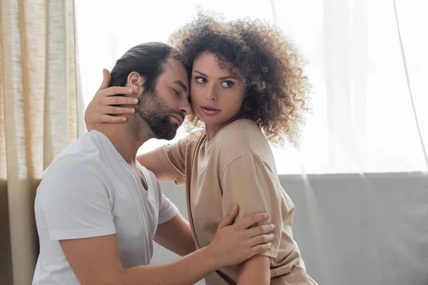 Curly woman in beige t-shirt hugging bearded boyfriend in bedroom - foto de stock