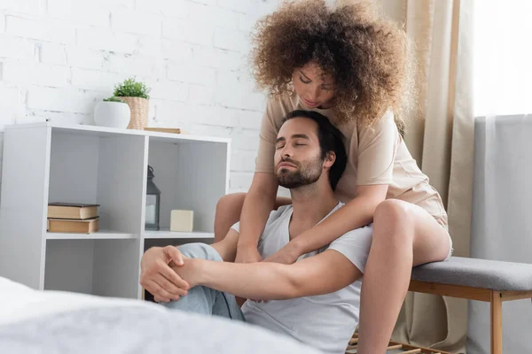 Mujer joven rizada sentada en la cama banco abrazando novio barbudo con los ojos cerrados en el dormitorio - foto de stock