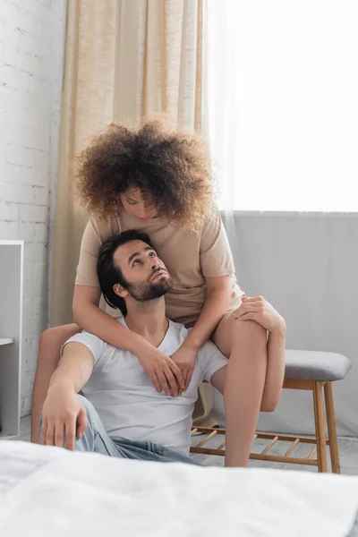 Curly woman sitting on bed bench hugging bearded boyfriend in bedroom — Stock Photo