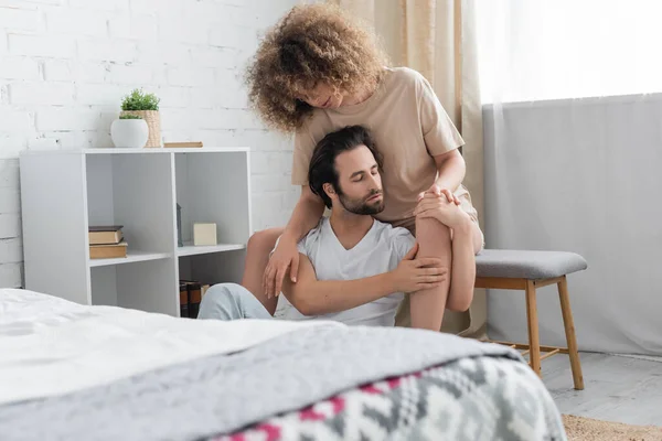 Curly woman sitting on bed bench hugging bearded boyfriend with closed eyes in bedroom - foto de stock