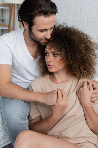Bearded man in jeans and white t-shirt sitting and hugging curly woman in bedroom - foto de stock