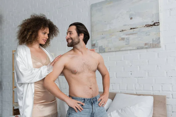 Сексуальная женщина в шелковом халате смотрит на бойфренда без рубашки в джинсах, позирующего с руками на бедрах — стоковое фото