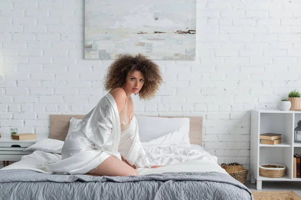 Кучерява і сексуальна молода жінка в білому шовковому халаті сидить на ліжку в сучасній спальні — стокове фото