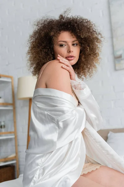 Curly and sexy young woman in white silk robe touching shoulder — Foto stock