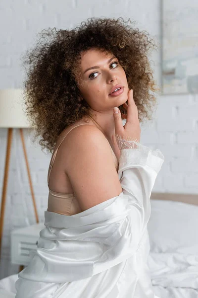Curly and sexy young woman in white silk robe touching face in bedroom — Foto stock