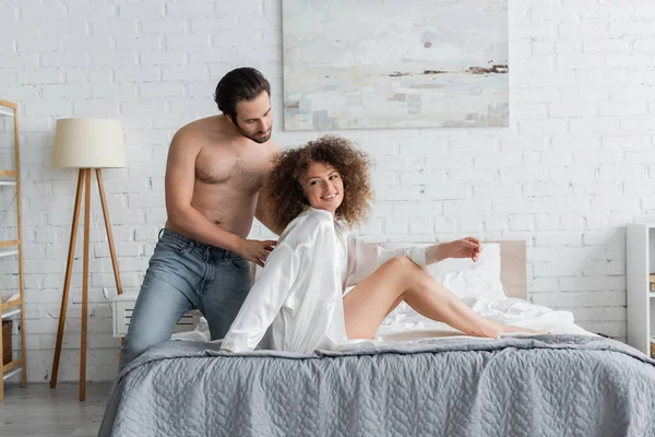 Shirtless man in jeans looking at cheerful and tattooed woman in bedroom — Stockfoto