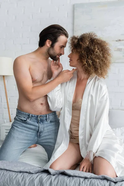 Shirtless man in jeans looking at curly woman in white satin robe — Stockfoto