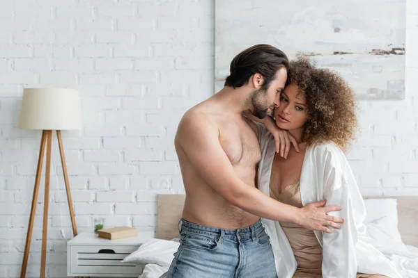 Shirtless man in jeans seducing curly woman in white satin robe - foto de stock